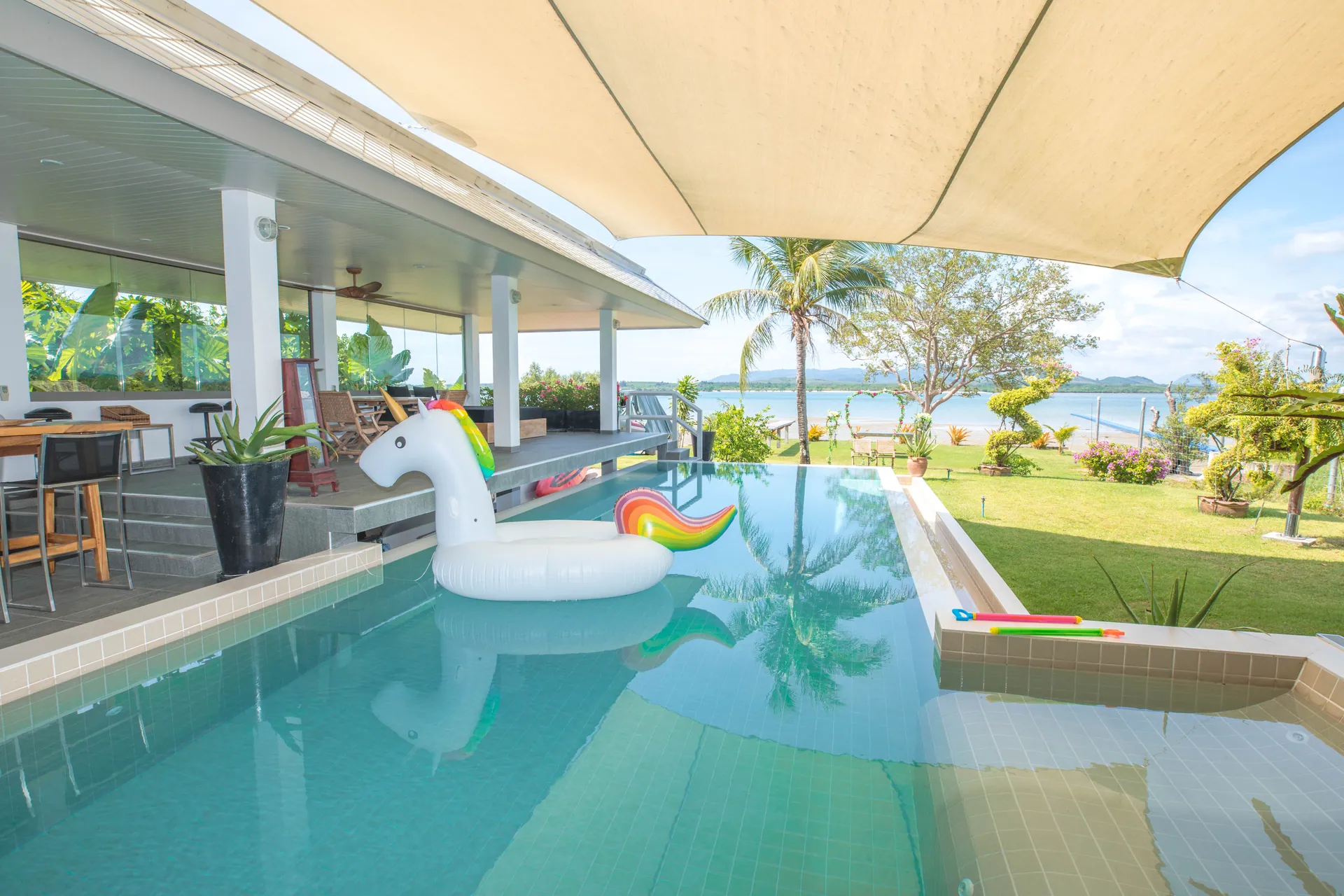 Garden pool under a shade sail, with an inflatable float and a lawn leading down to the sea