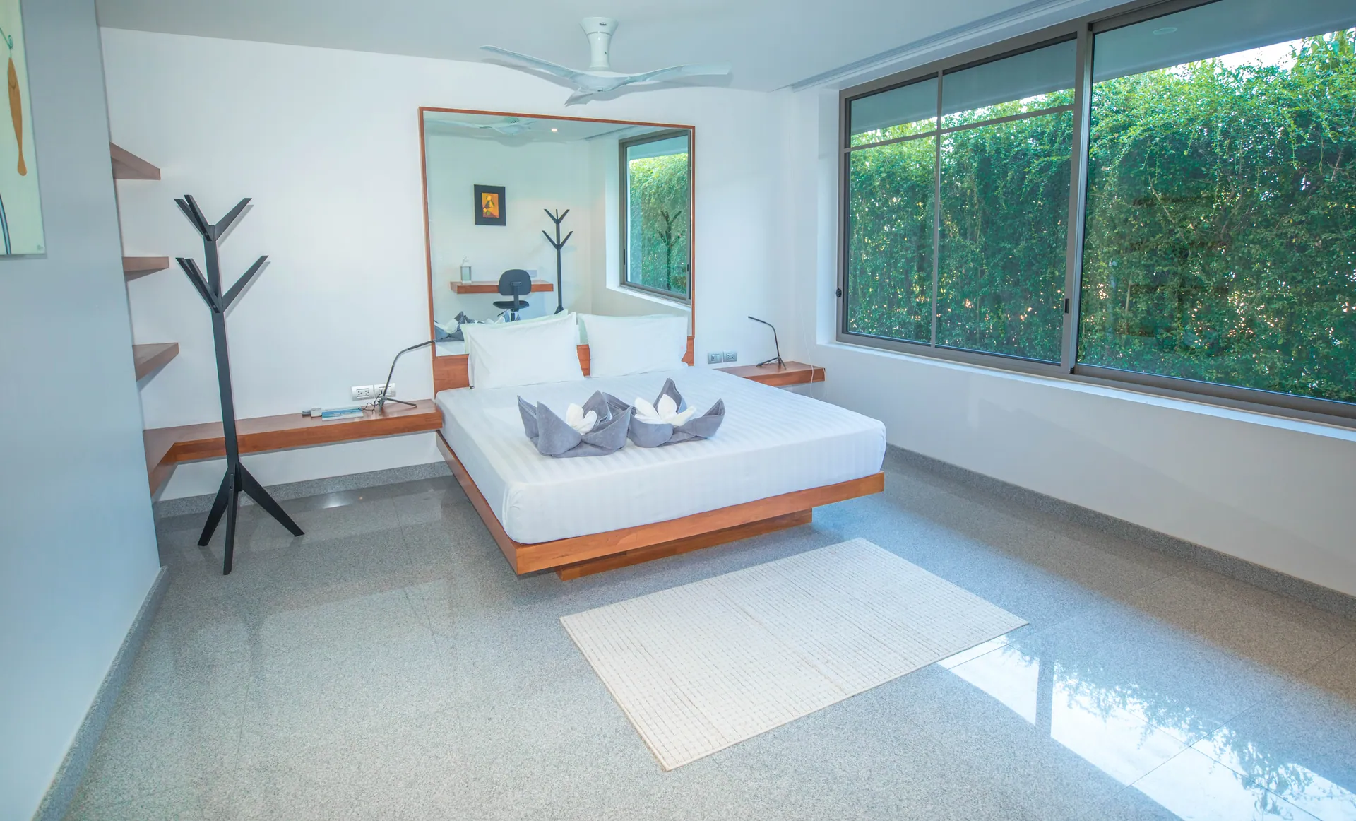 Bedroom with a platform bed, a coat stand, a wall mirror and wide windows onto a green hedge