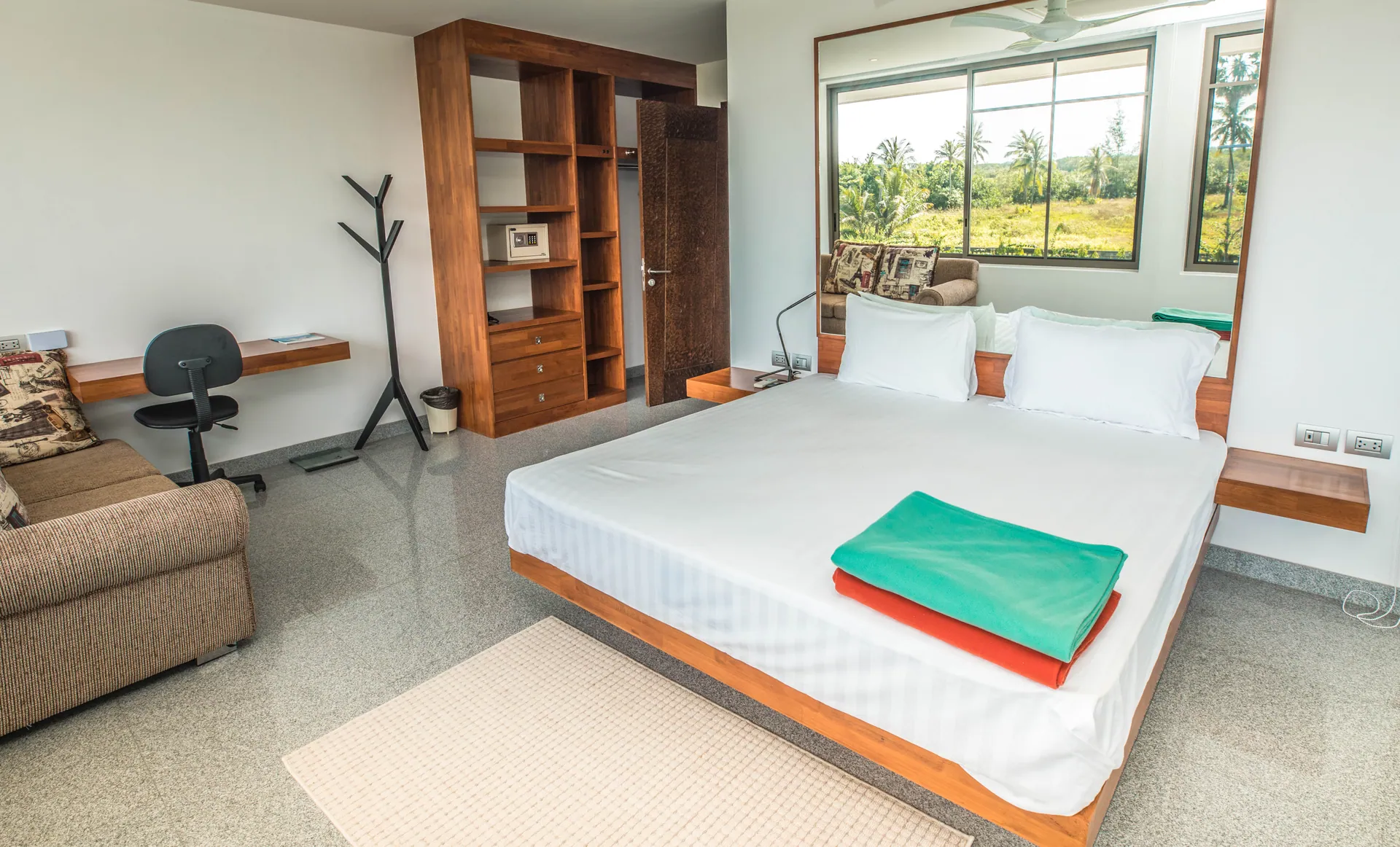 Bedroom with a platform bed, a sofa, a desk and open timber shelving, with windows onto palms and fields