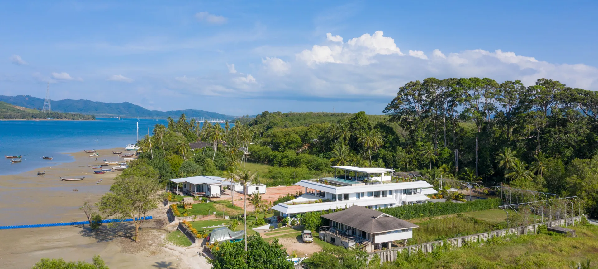 Aerial view of the shoreline, with longtail boats on the sand flats, the villa among trees and hills across the water