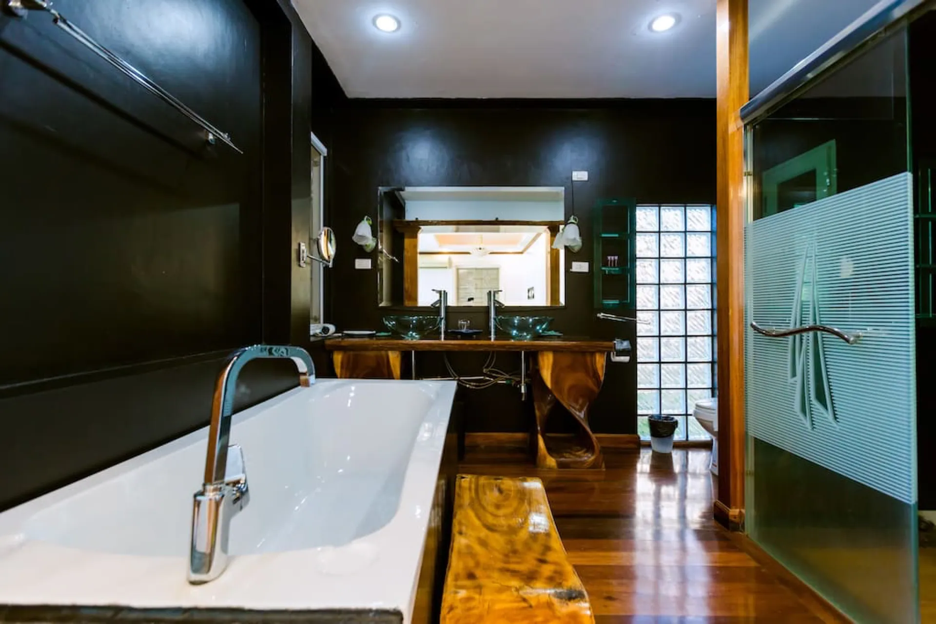 Dark-walled bathroom with a bathtub, twin basins on a timber vanity and a glass shower
