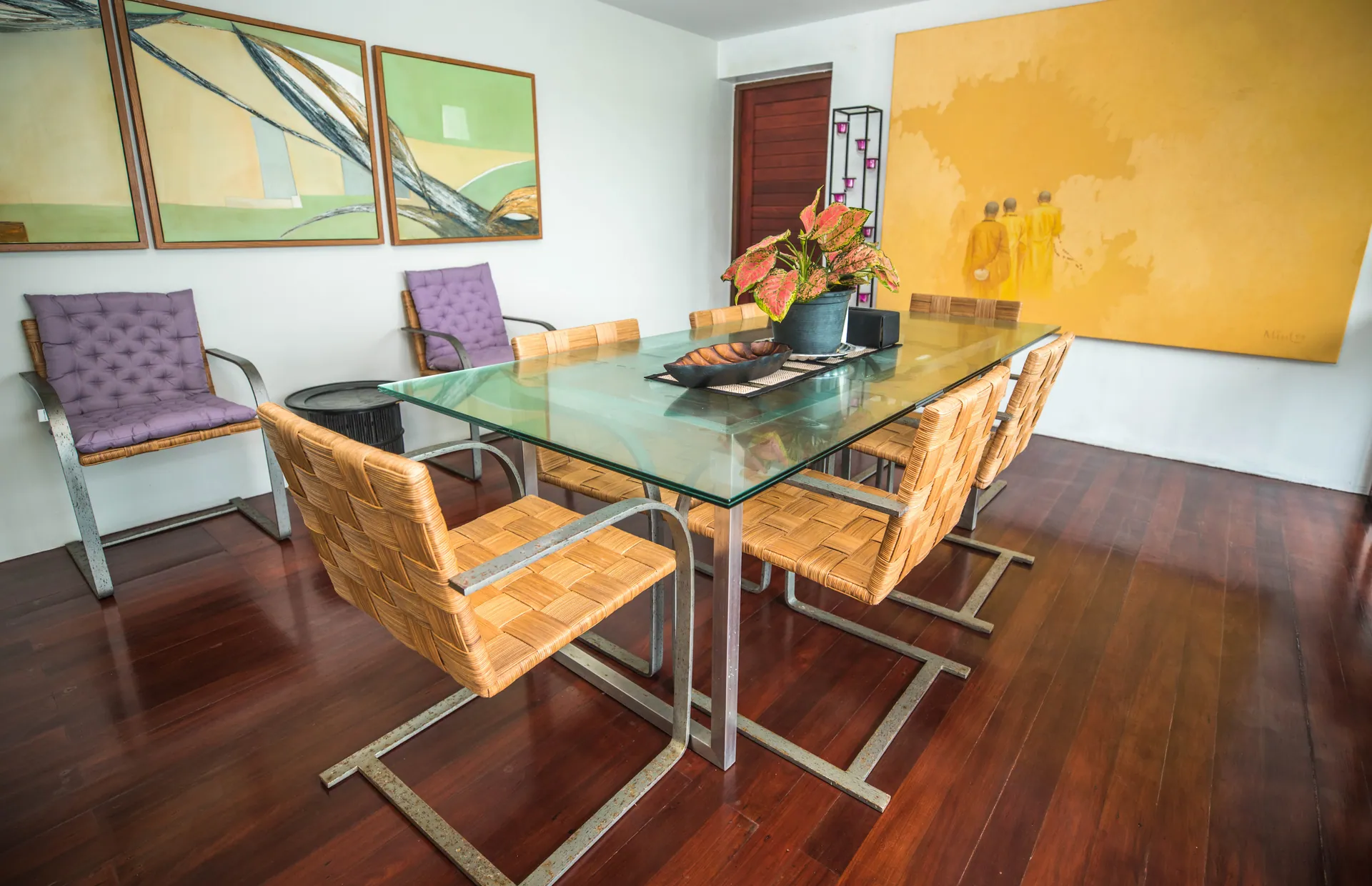 Glass dining table with woven chairs, beside a large yellow painting