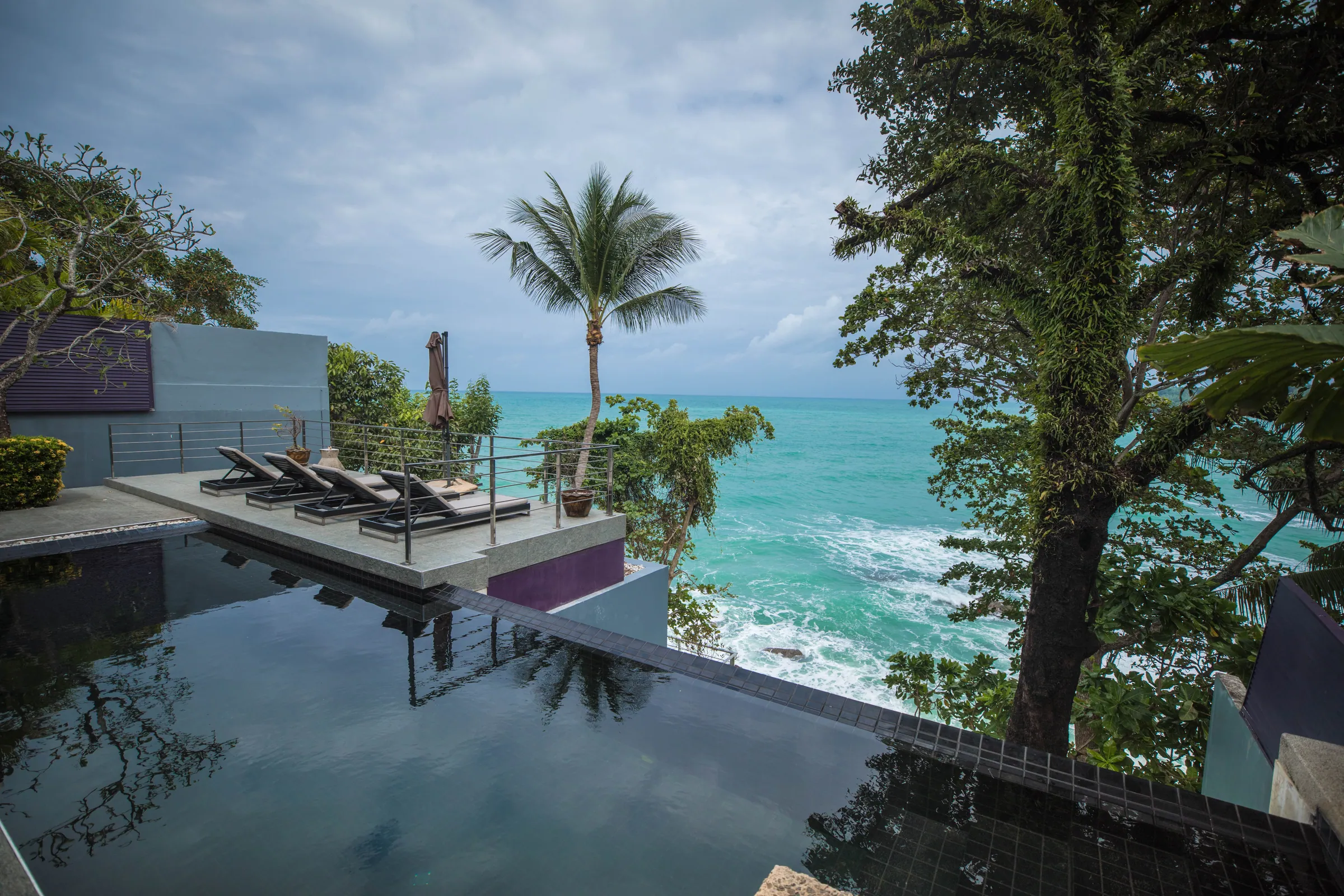Infinity-edge pool on a terrace above the sea, with sun loungers and a palm tree against the sky