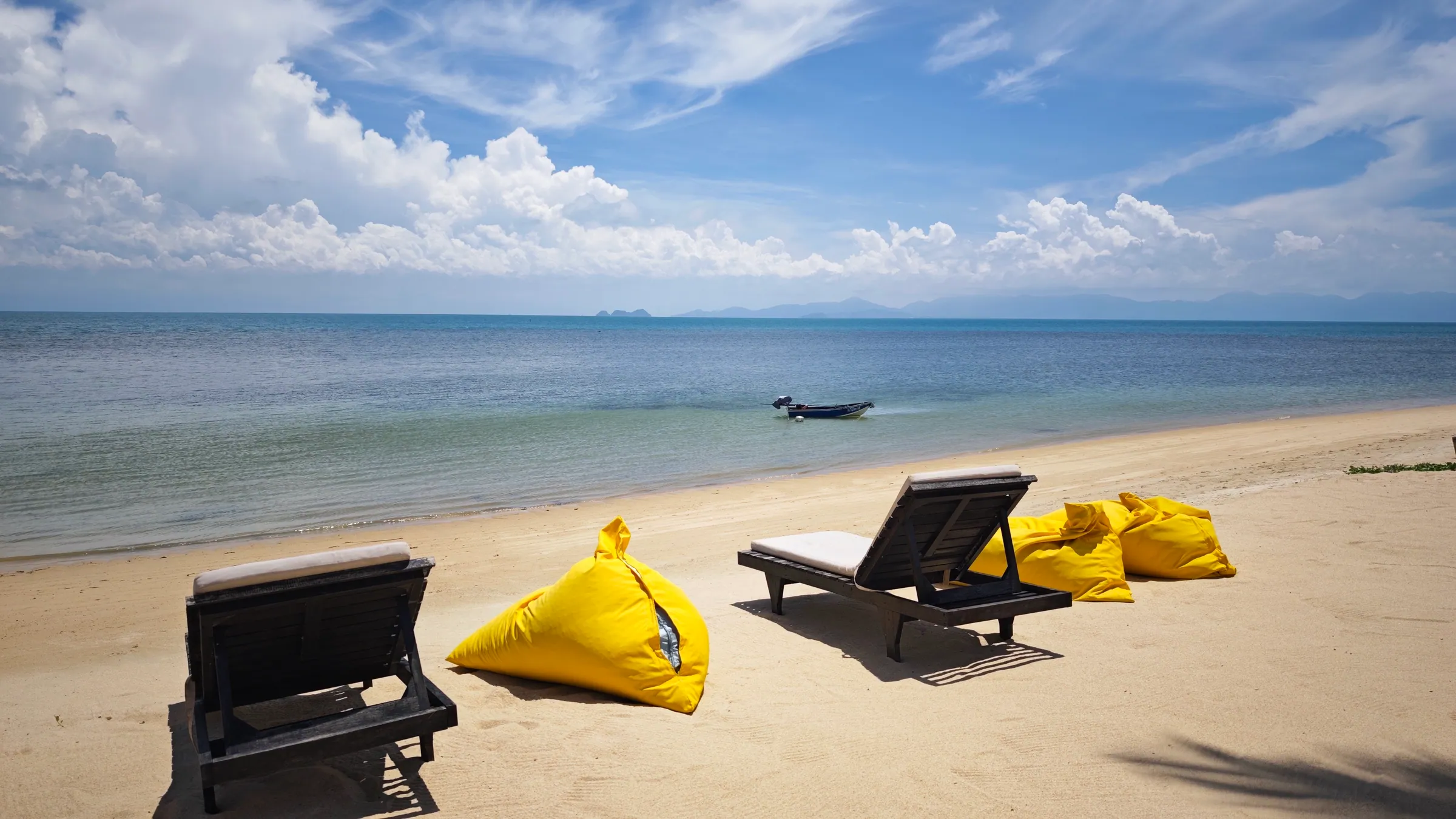 Sun loungers and yellow bean bags on the beach in front of the villa, with calm sea and distant islands under a blue sky