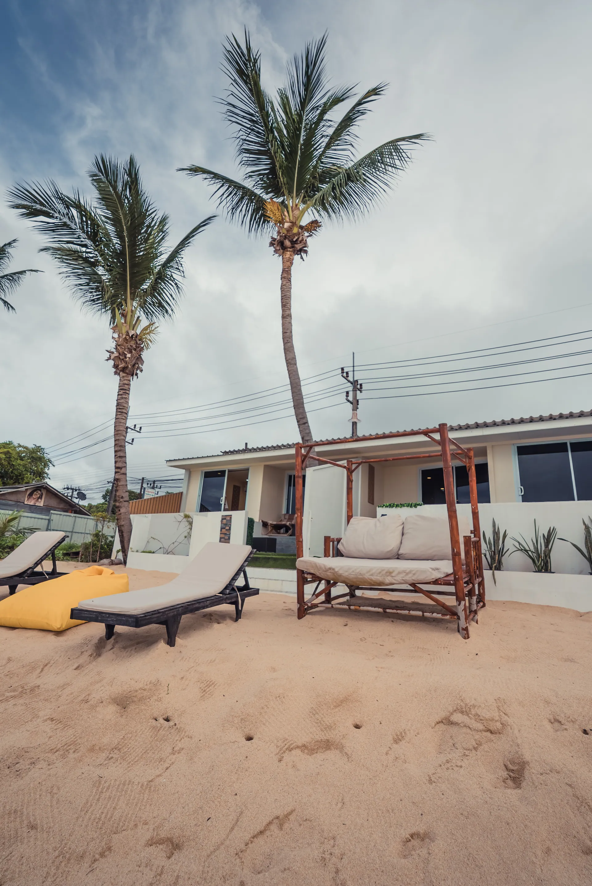 Timber daybed and a lounger on the sand in front of the villa, framed by tall coconut palms