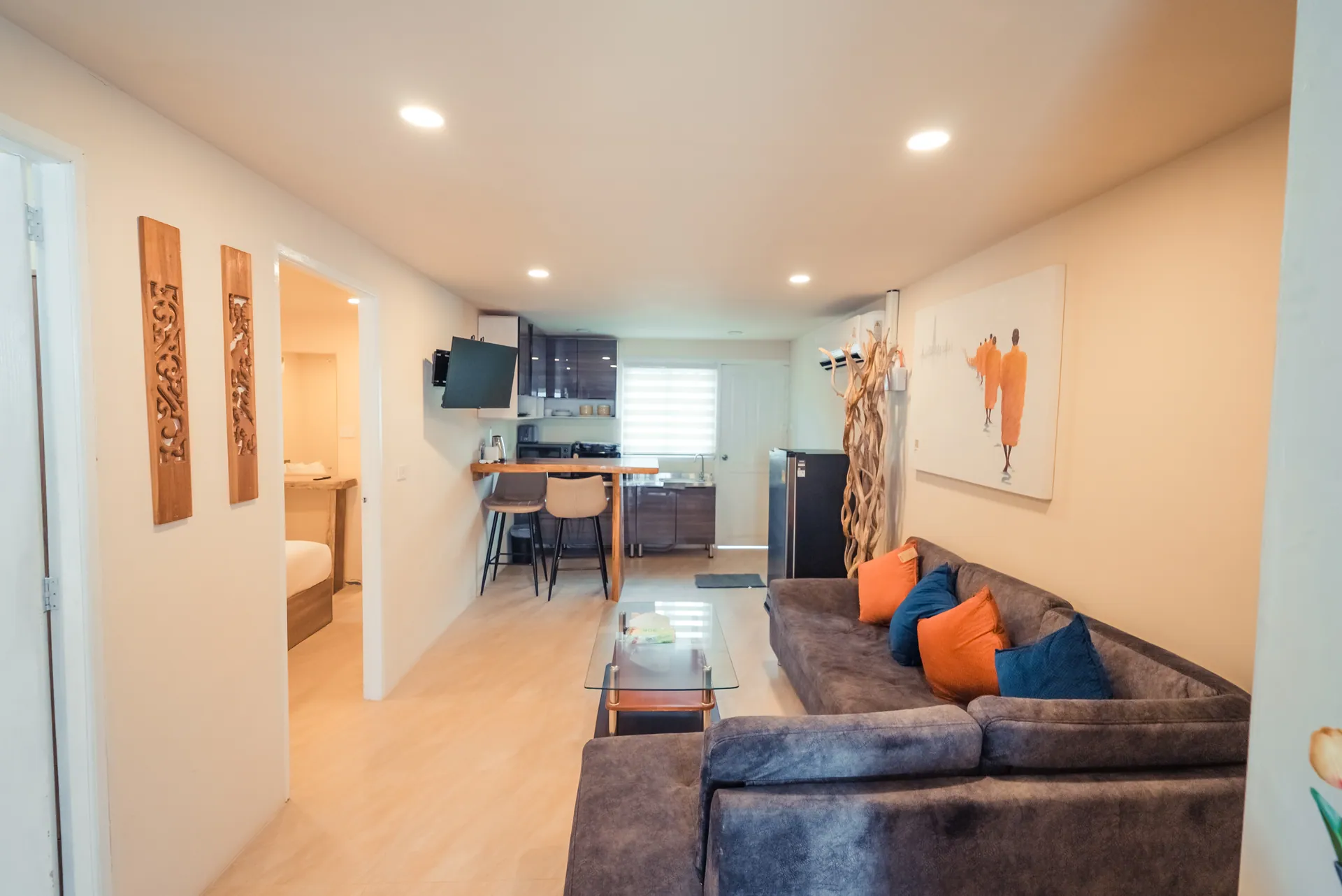 Living room with a grey corner sofa and orange and blue cushions, opening through to a kitchenette with a breakfast bar