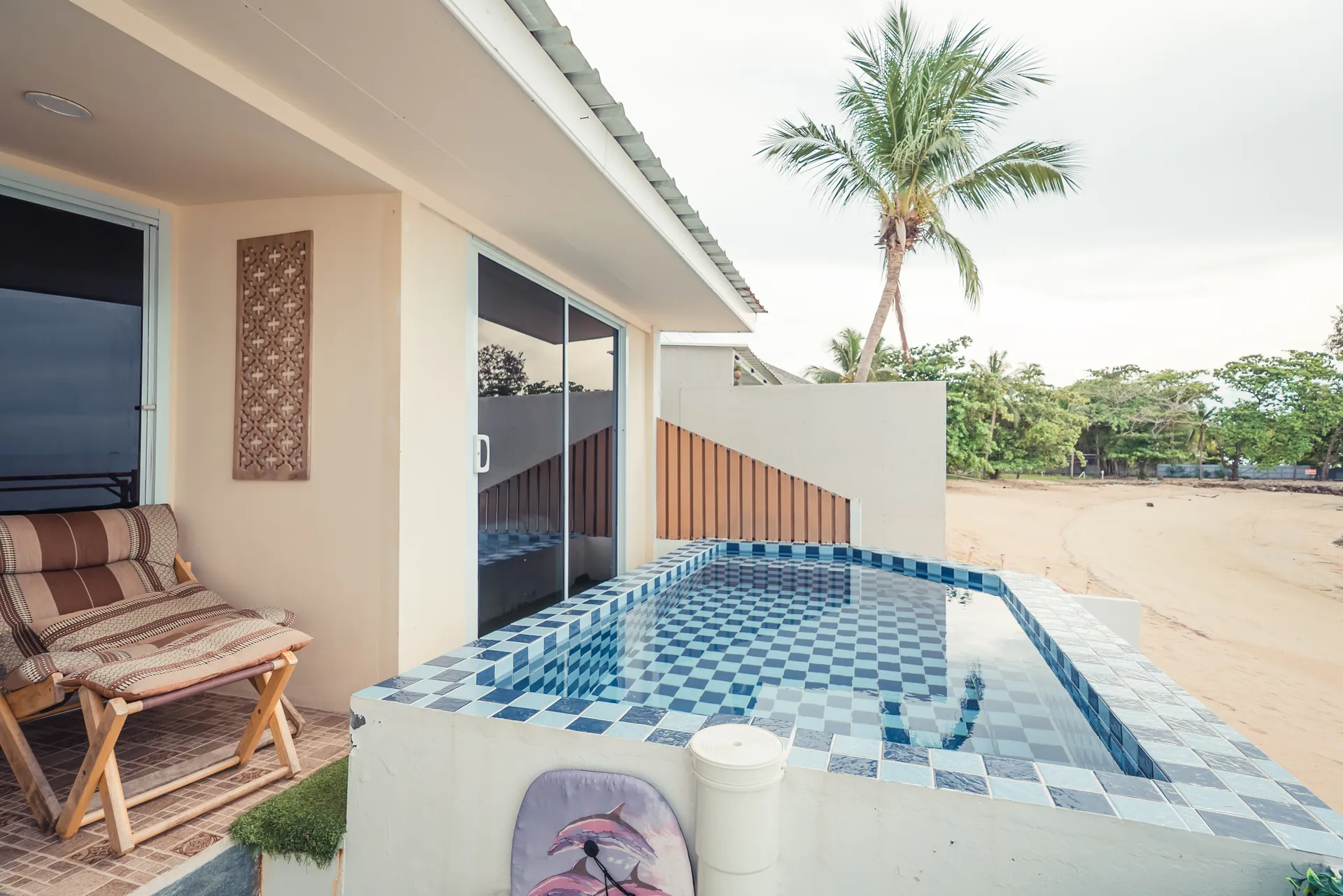 Blue mosaic-tiled plunge pool beside a covered terrace with a lounge chair, with a palm and the beach beyond