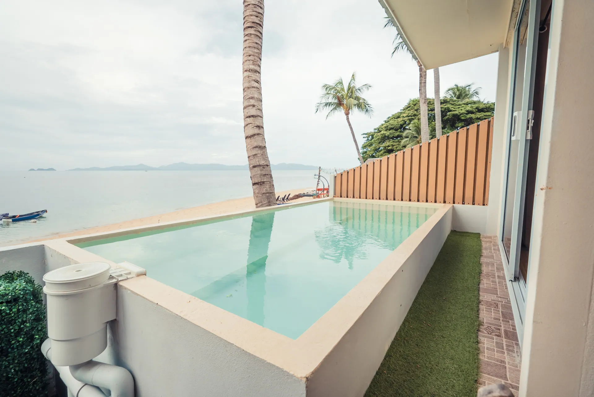 Private plunge pool on a terrace with artificial turf, looking past a palm trunk to the beach and the sea
