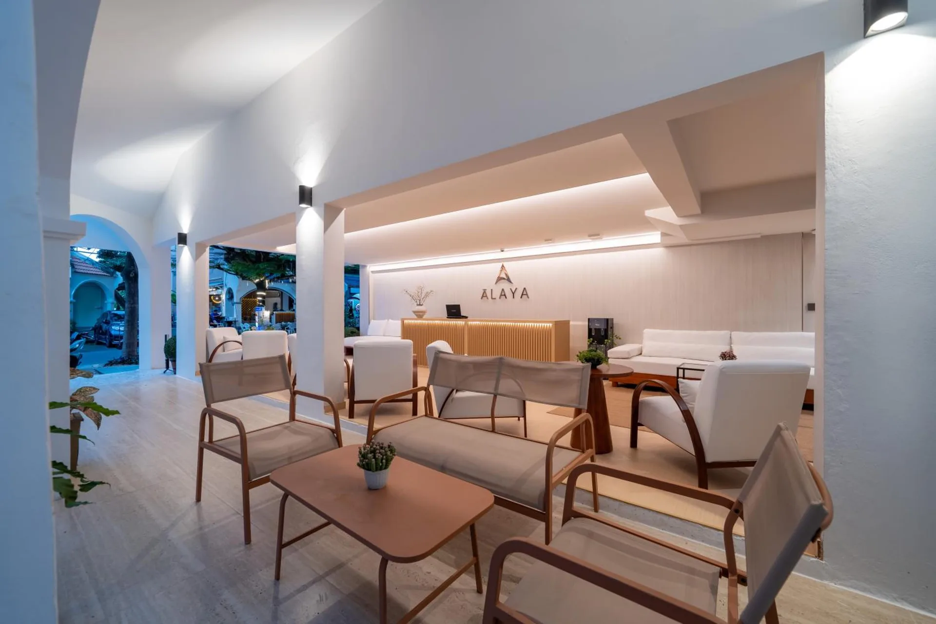 Lobby lounge beneath white arches, with timber armchairs and the Alaya logo behind the front desk